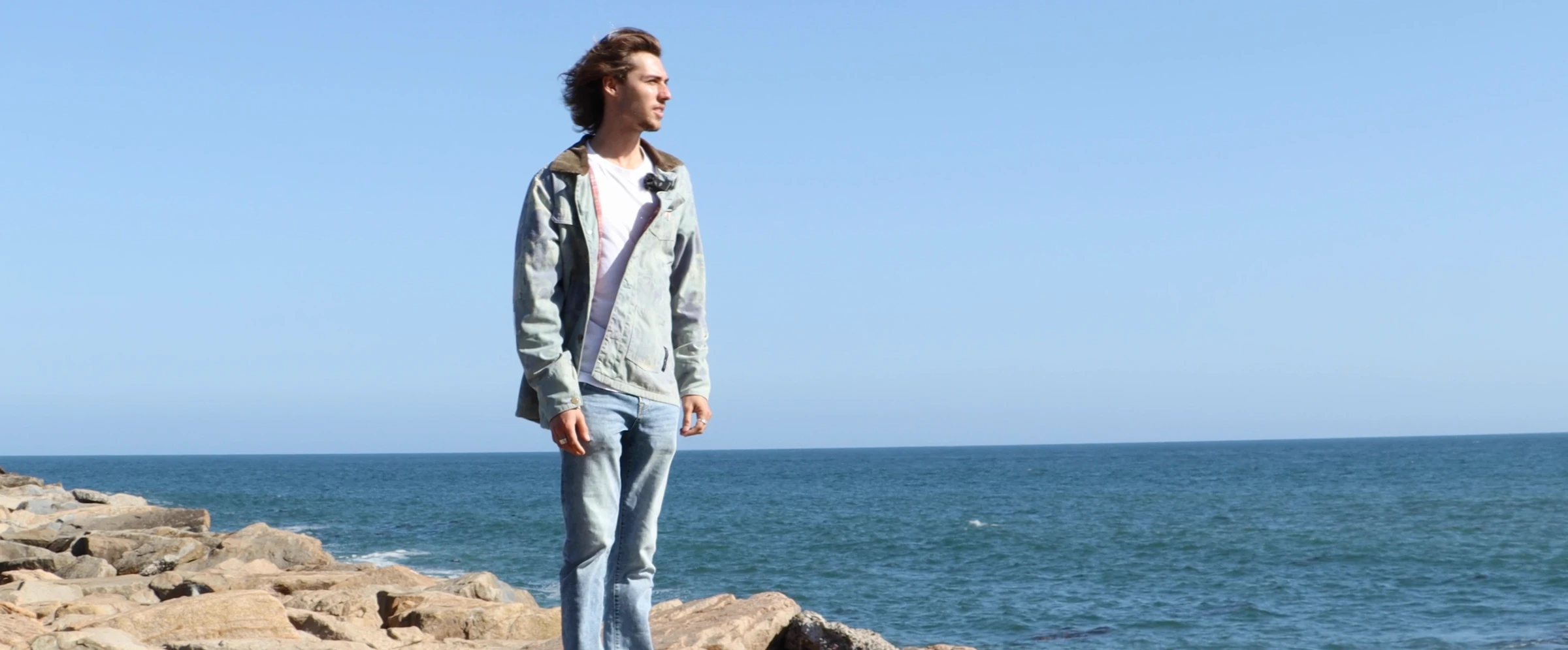 Will Jandro stands on coastal rocks above the ocean, hair blowing in the wind