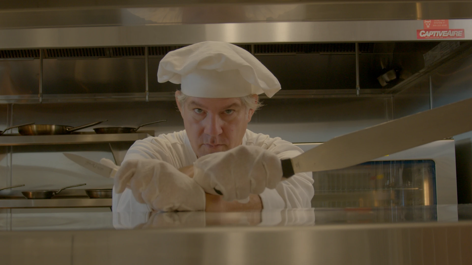 A chef in a white toque stares down the lens, arms crossed, holding two knives